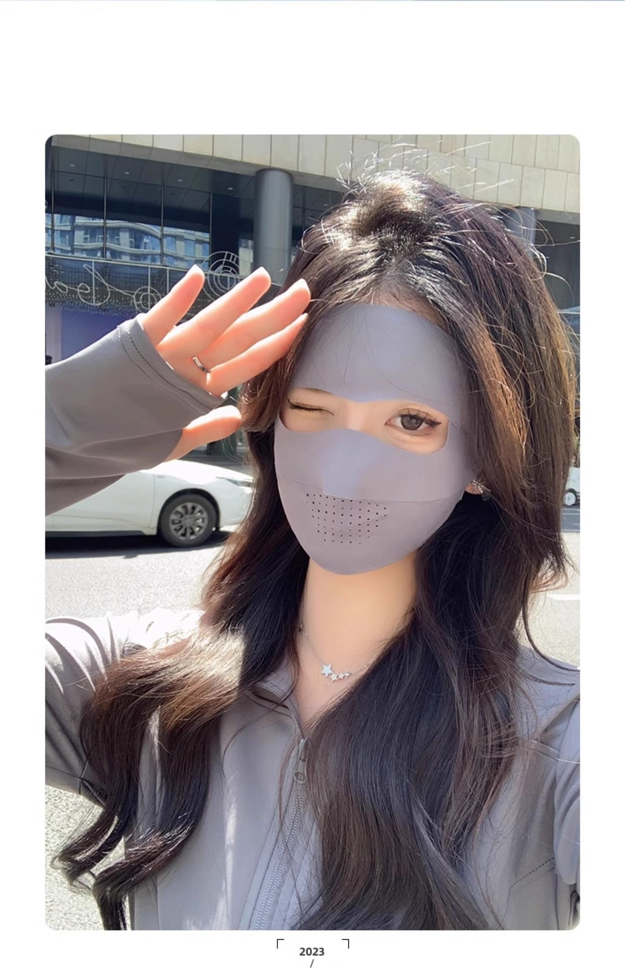 A person with long, wavy hair is taking a selfie outdoors, wearing a Facekini Full Face Sunscreen Mask in gray. The mask features cutouts over the eyes and mouth. Using one hand as a sunshade while winking, they capture buildings and a street in the background.