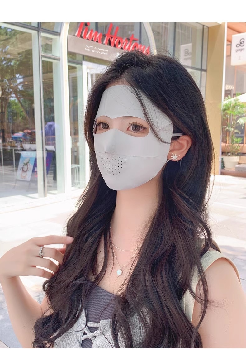 A woman is wearing a Facekini Full Face Sunscreen Mask made of breathable thin ice silk, which covers her head and nose with holes for breathing. She has long black hair and is wearing a sleeveless gray top. In the background, there is a Tim Hortons cafe with glass windows.