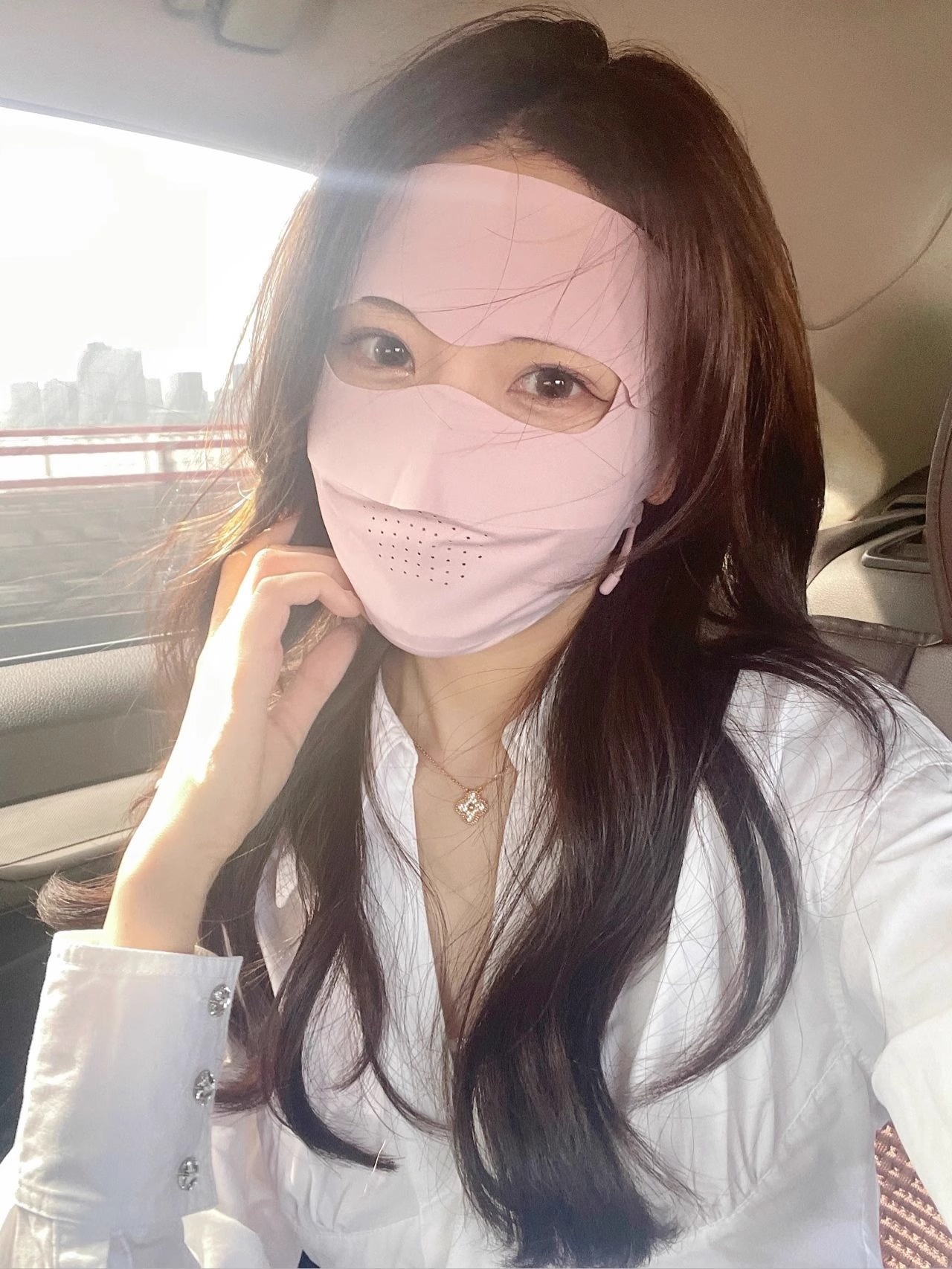 A woman with long brown hair sits in a car, wearing a white shirt and a Facekini Full Face Sunscreen Mask in light pink ice silk that covers her mouth and nose. Sunlight streams in through the window as she casually rests her hand on her chin while looking at the camera.