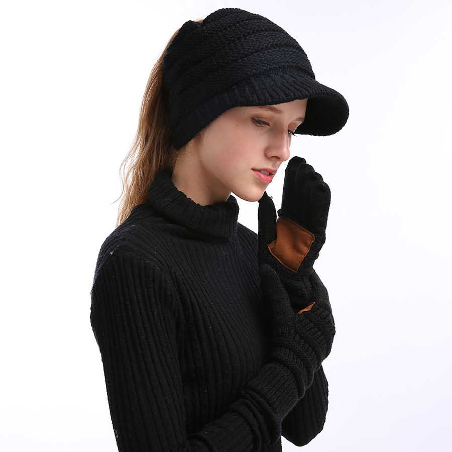 A woman stands against a white background, wearing a black knit hat and sweater perfect for winter. Her long hair is tied back, and she thoughtfully touches her face with European And American Wool Knitted Turn-over Labeling Touch Screen Gloves, which feature distinctive black gloves with brown palms. The ensemble is completed with cozy textures like acrylic fiber to combat the chill.